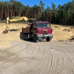 Żółta koparka nabiera piasek do wywrotki Tatra na tle lasu sosnowego i wykopu w słoneczny dzień.