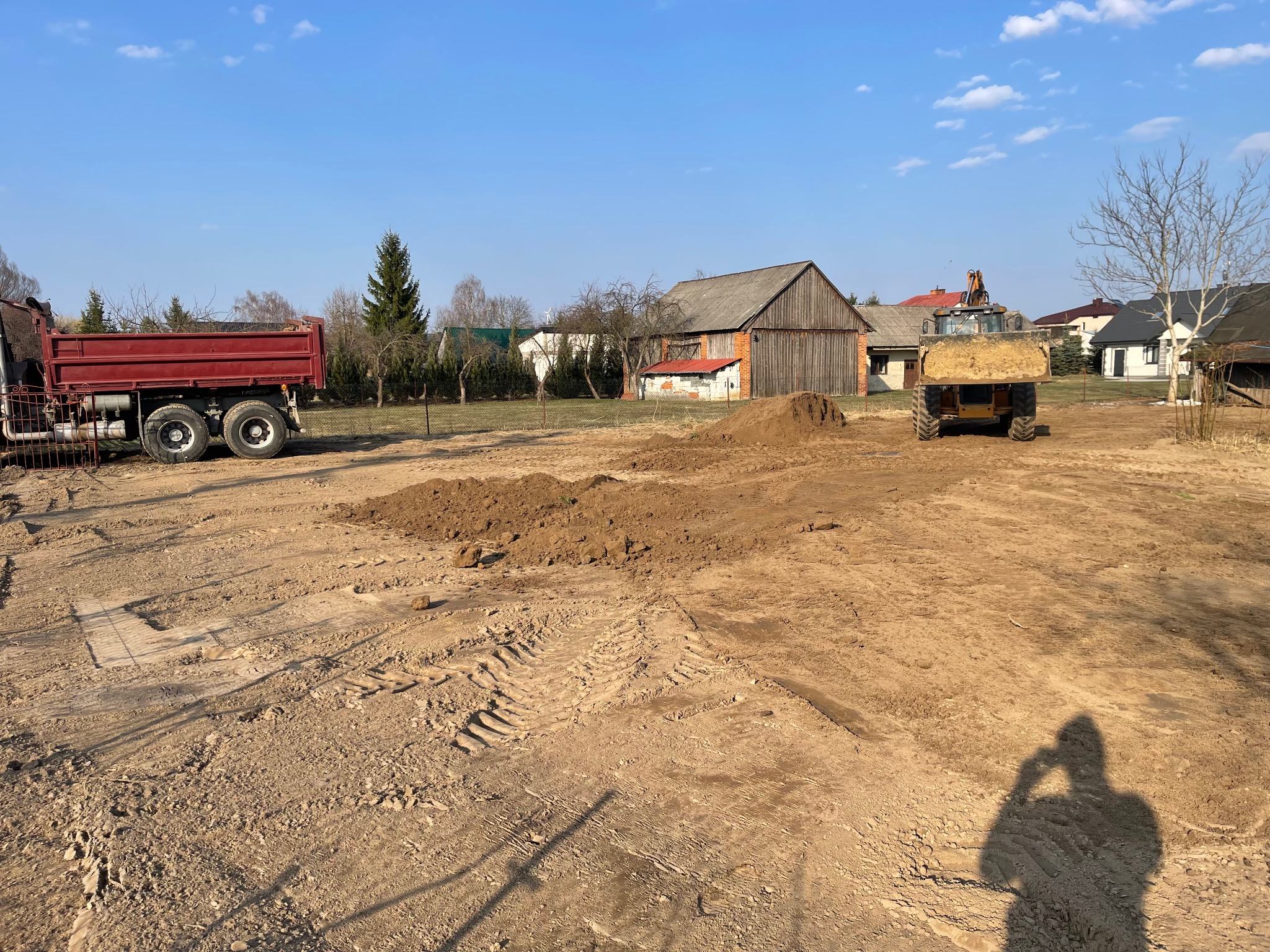 Ciężarówka wywrotka i ładowarka na placu budowy z pryzmą ziemi, w tle budynki gospodarcze i domy jednorodzinne.