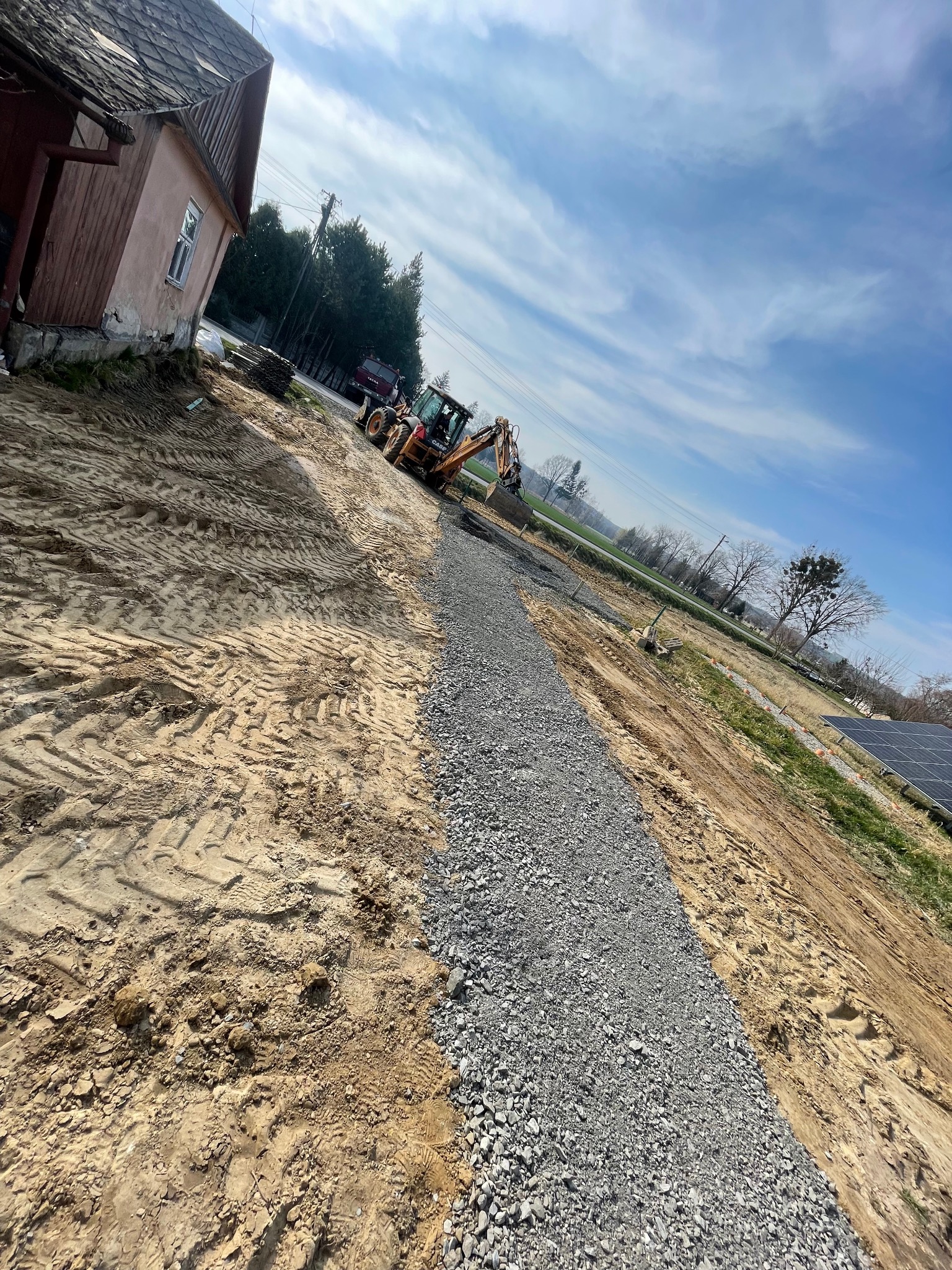 Żółta koparko-ładowarka CASE wyrównuje wąski pas żwiru na piaszczystym terenie budowy, obok budynku z czerwonymi elementami, w tle widoczne drzewa i niebieskie niebo z chmurami.