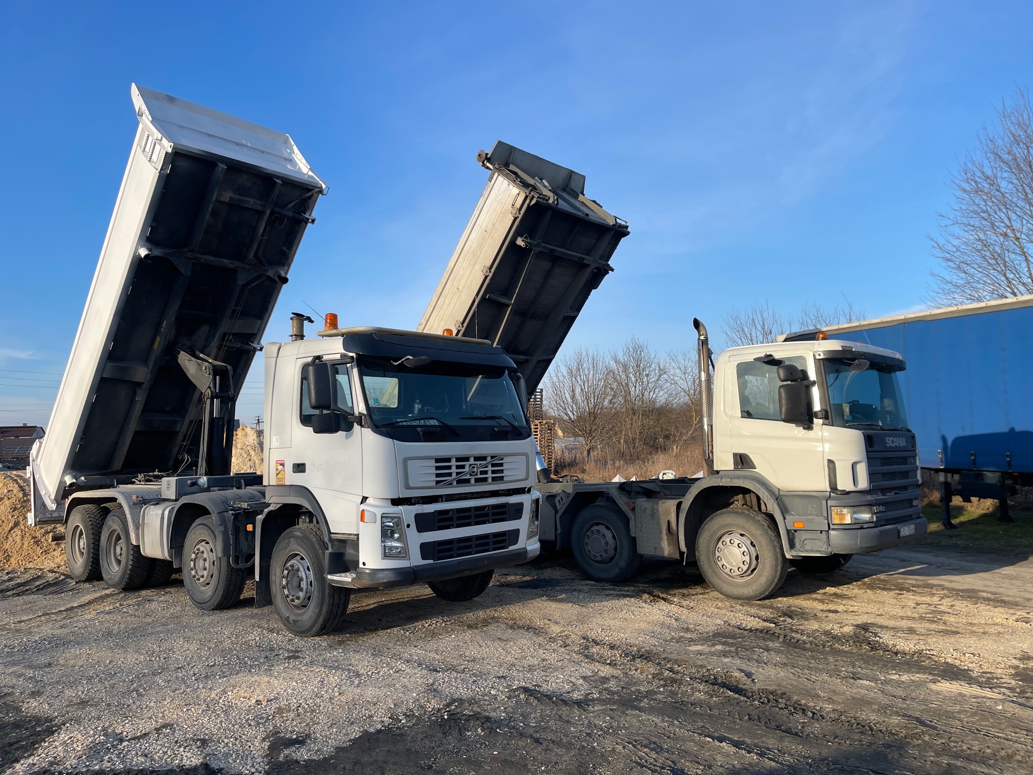 Dwa białe samochody ciężarowe typu wywrotka z podniesionymi skrzyniami ładunkowymi na żwirowym placu w słoneczny dzień.