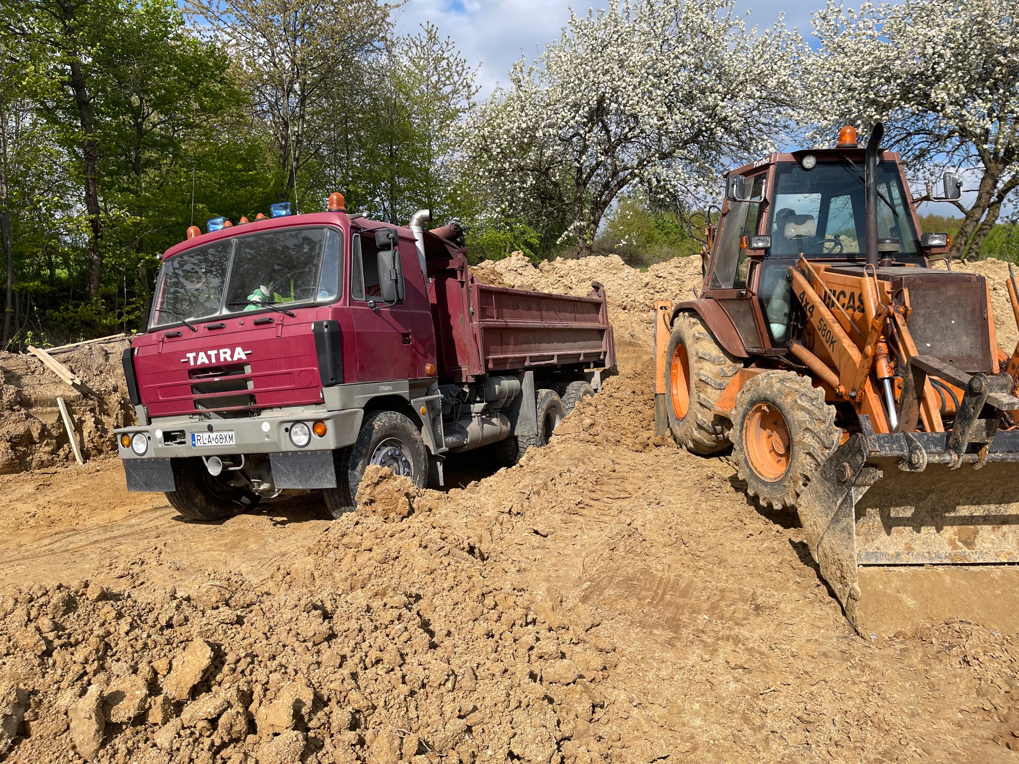 Czerwona ciężarówka Tatra i pomarańczowa koparko-ładowarka na placu budowy, w tle kwitnące drzewa.