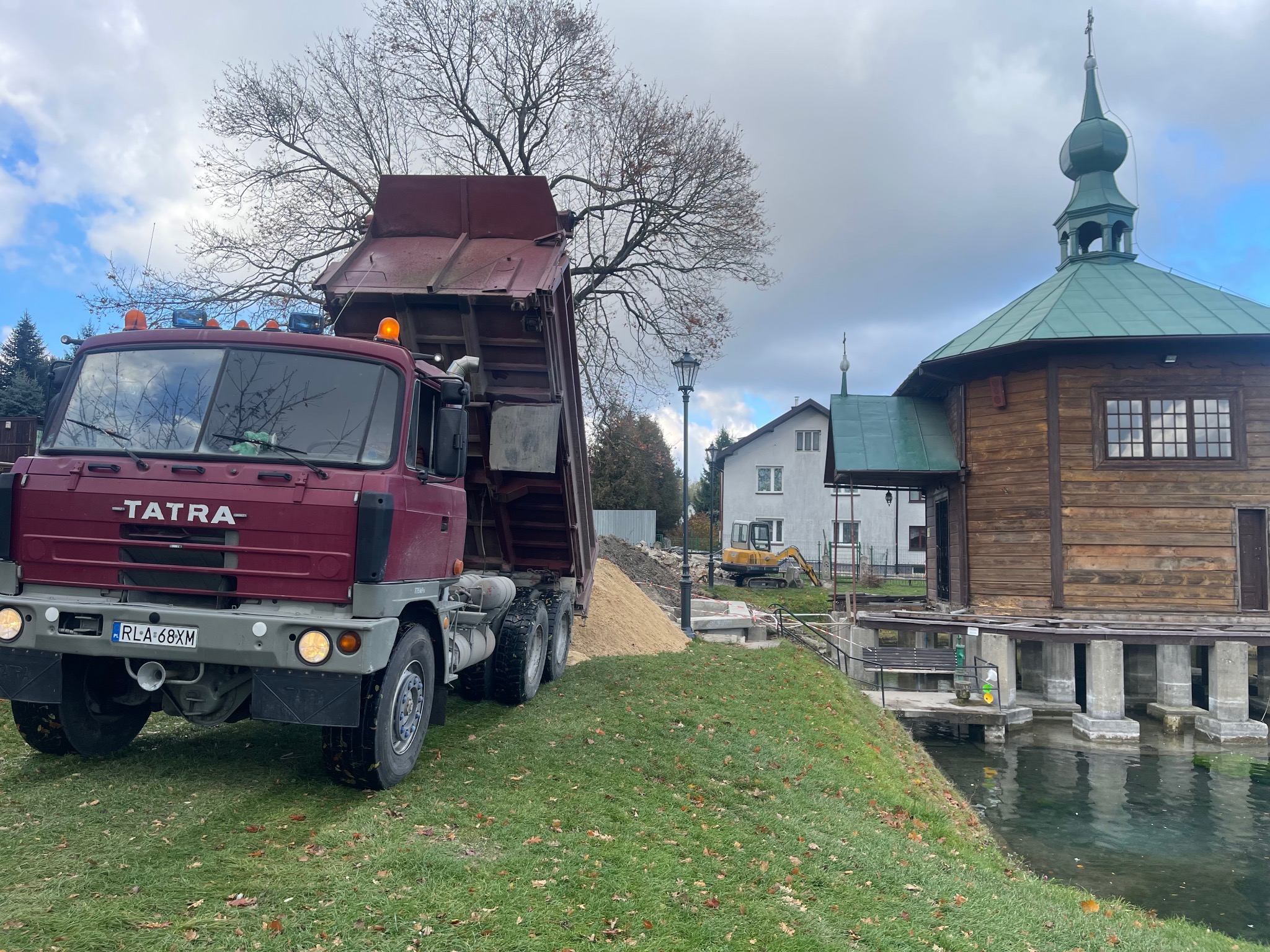Ciężarówka Tatra z podniesioną skrzynią wyładowuje piasek na trawniku obok drewnianego budynku z zielonym dachem, w tle widoczna koparka.