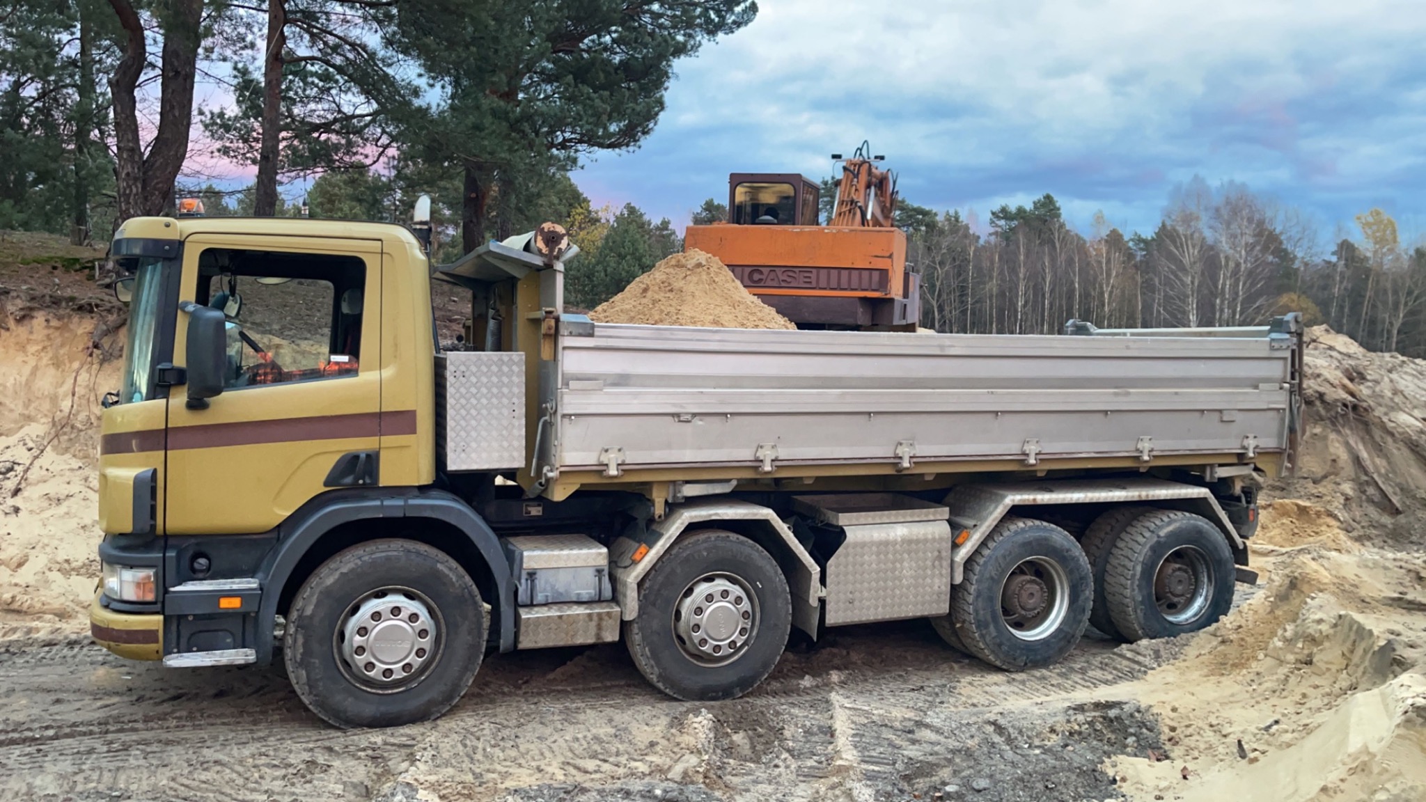 Żółta ciężarówka z ładunkiem piasku na mulistej ziemi, w tle koparka i drzewa, pochmurne niebo.
