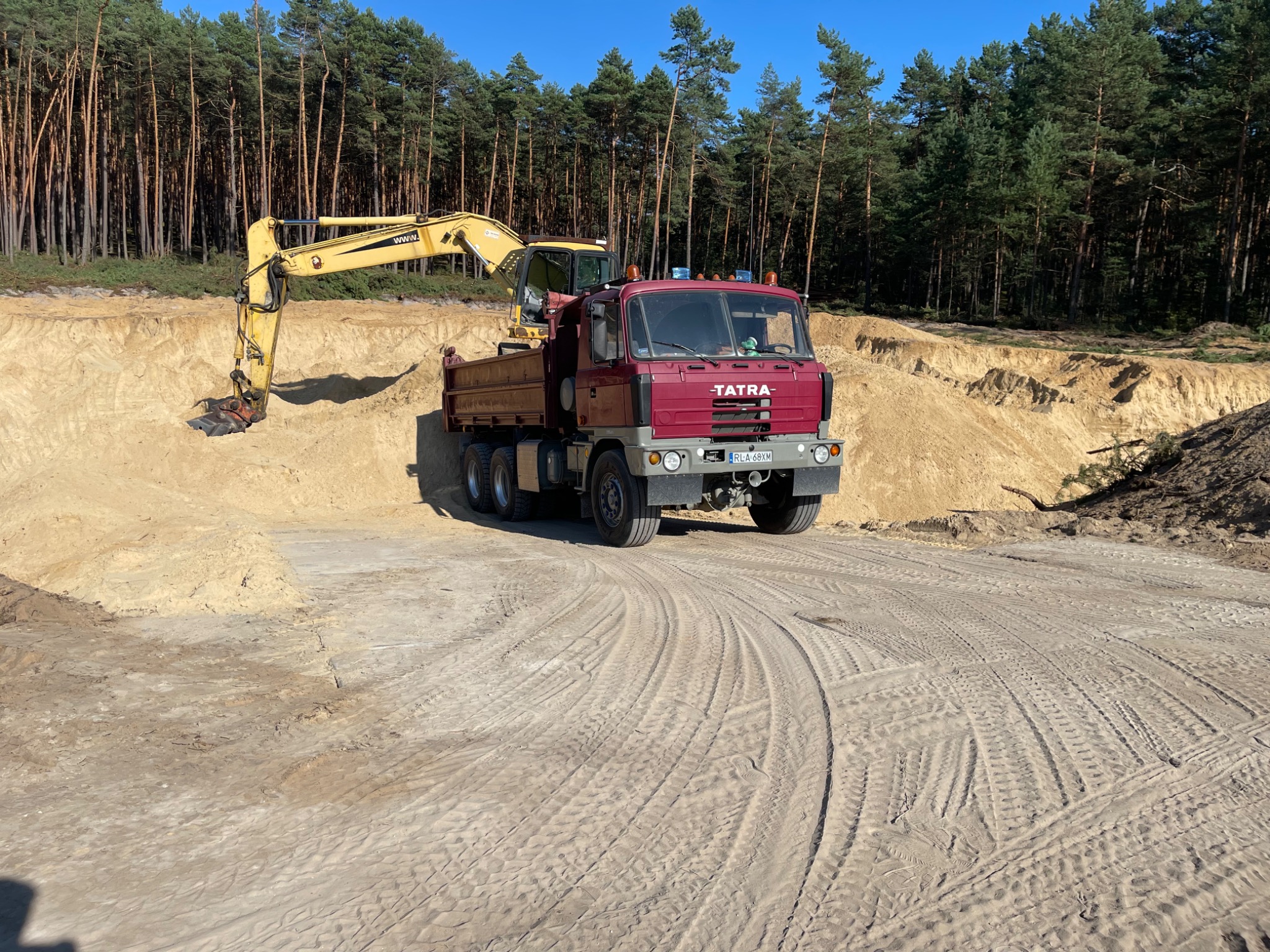 Żółta koparka nabiera piasek do wywrotki Tatra na tle lasu sosnowego i wykopu w słoneczny dzień.