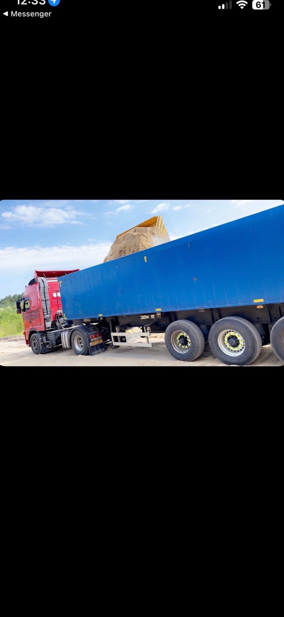 Czerwona ciężarówka z niebieską naczepą wypełniona piaskiem, widoczna na tle błękitnego nieba z lekkimi chmurami.