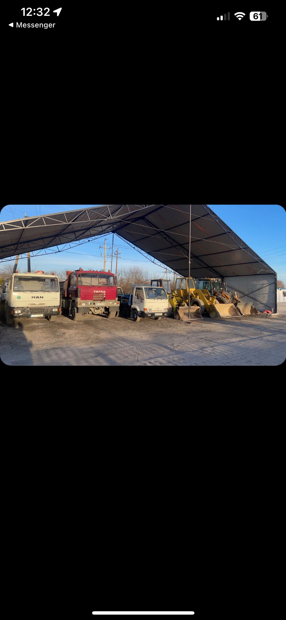 Rząd pojazdów budowlanych: ciężarówki MAN i Tatra oraz koparki, zaparkowane pod zadaszeniem w słoneczny dzień.