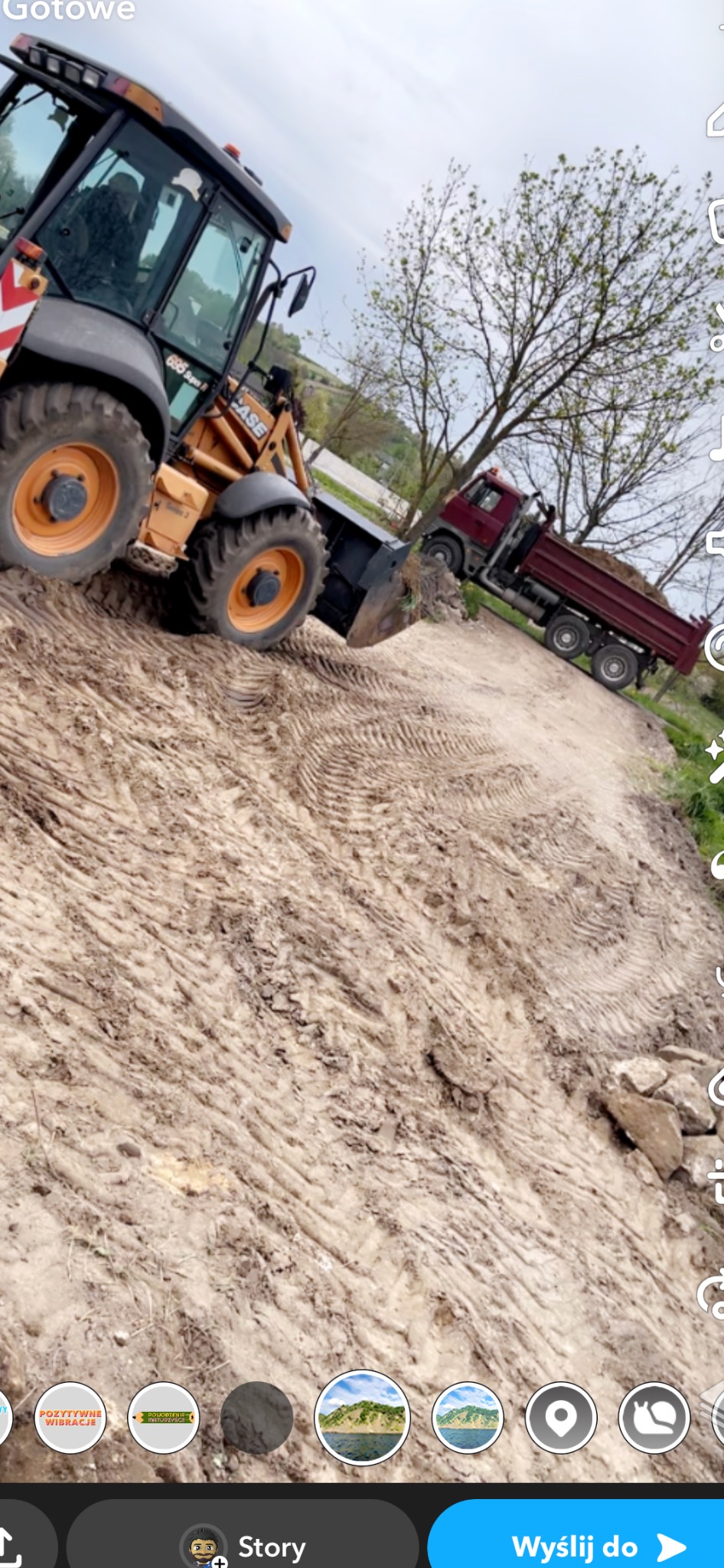 Żółto-czarna koparko-ładowarka na nierównym, brązowym terenie z widocznymi śladami opon, w tle czerwona wywrotka i drzewa pod pochmurnym niebem.