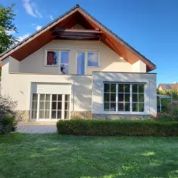 Jasny, dwukondygnacyjny dom z beżową elewacją, brązowym dachem i białymi oknami, otoczony zielonym ogrodem z kwitnącymi krzewami i drzewami w słoneczny dzień.