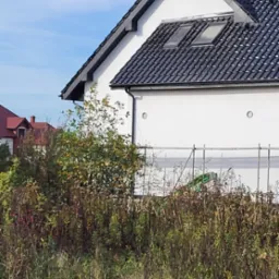 Boczny widok domu z białą elewacją, czarnym dachem z oknami dachowymi, widoczne rusztowania w tle, zarośnięte pole na pierwszym planie.