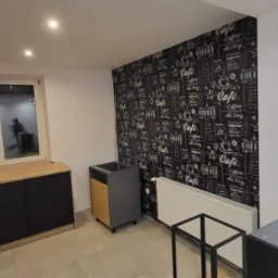 Wnętrze pokoju w trakcie wykańczania, z meblami kuchennymi w zabudowie, grzejnikiem i ścianą z tapetą w motywy kawowe, widoczne narzędzia na blacie.