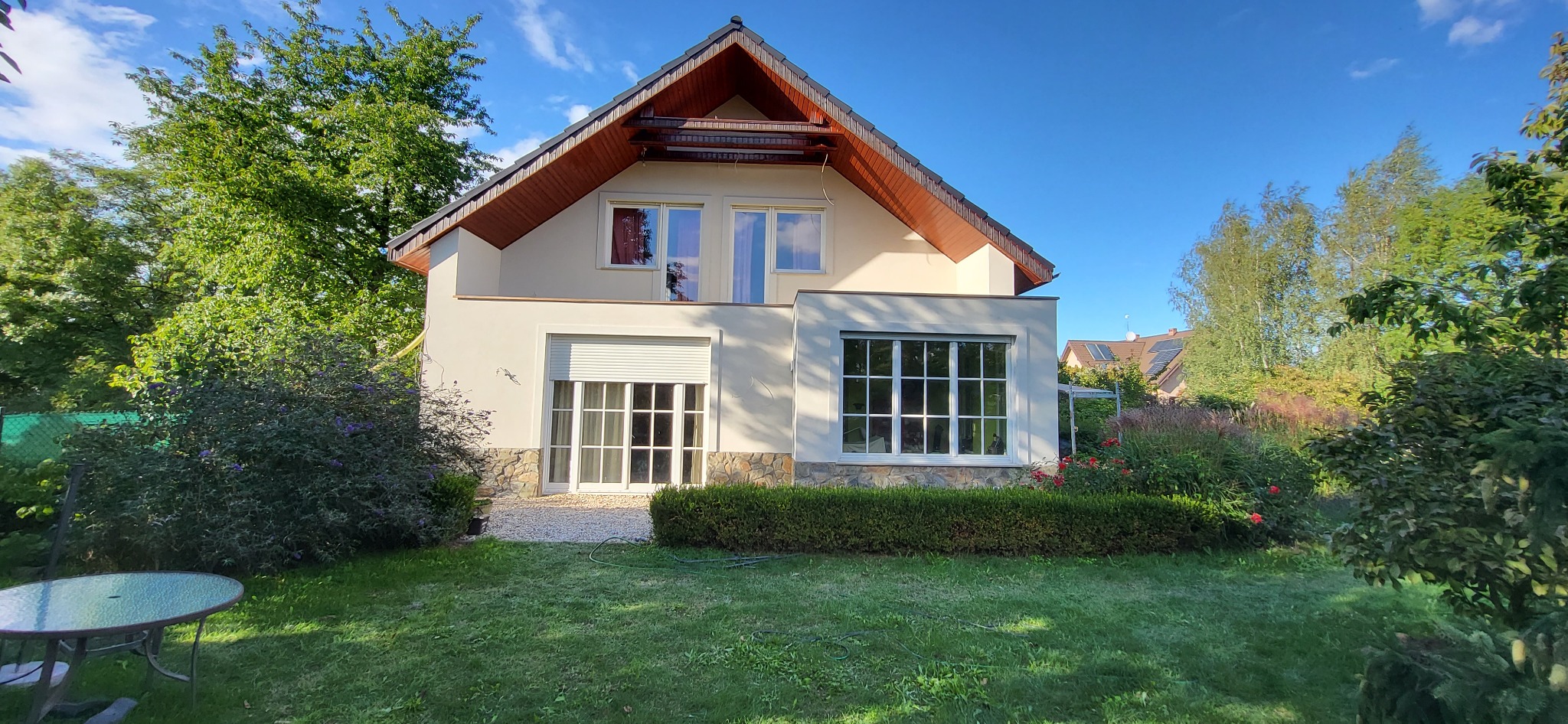 Jasny, dwukondygnacyjny dom z beżową elewacją, brązowym dachem i białymi oknami, otoczony zielonym ogrodem z kwitnącymi krzewami i drzewami w słoneczny dzień.