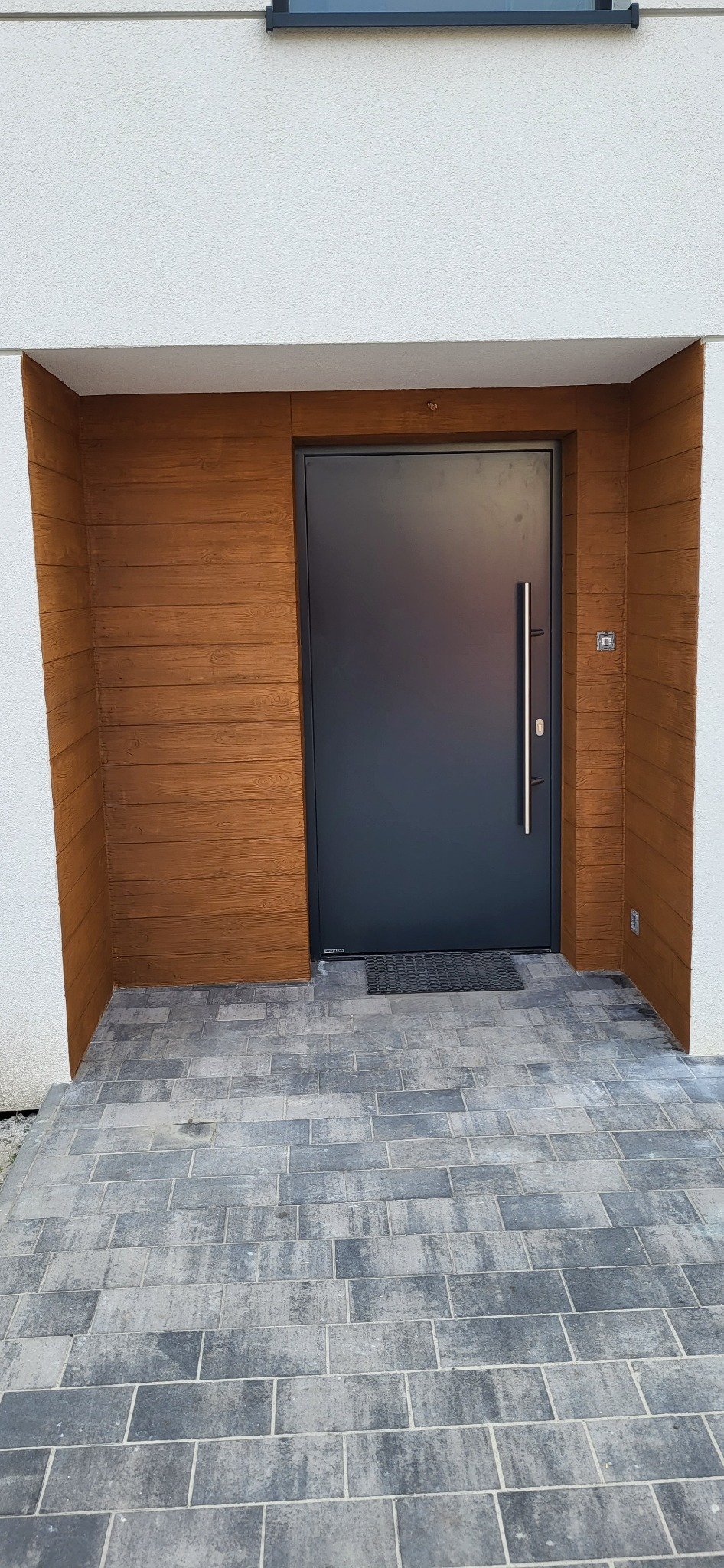Nowoczesne wejście do domu z szarą, minimalistyczną stolarką drzwiową, okładziną elewacyjną imitującą drewno i brukowanym podjazdem z szarej kostki.