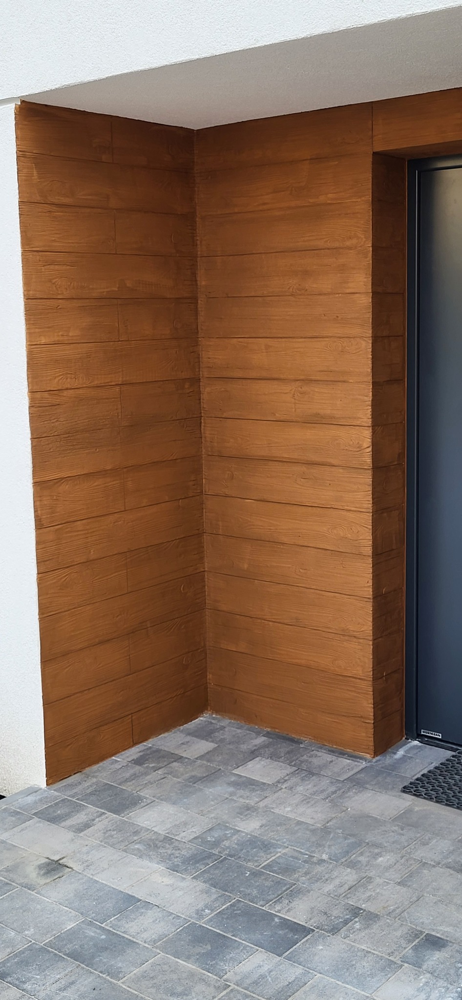 Elewacja budynku wykończona brązowymi panelami imitującymi drewno, z widocznym narożnikiem i fragmentem drzwi wejściowych, położona na szarej kostce brukowej.