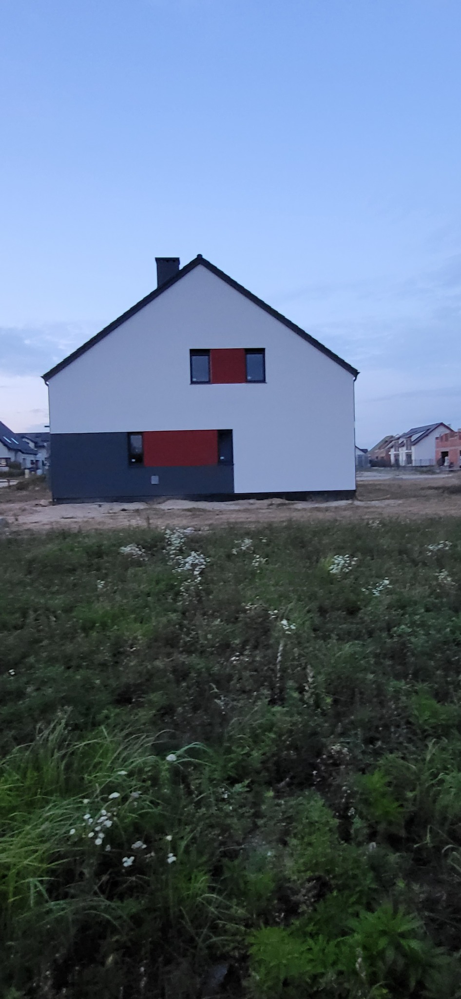 Nowoczesny dom jednorodzinny z białą elewacją i szarym cokołem, akcentowany czerwonymi panelami wokół okien, na tle zachmurzonego nieba i niezagospodarowanego terenu z roślinnością.