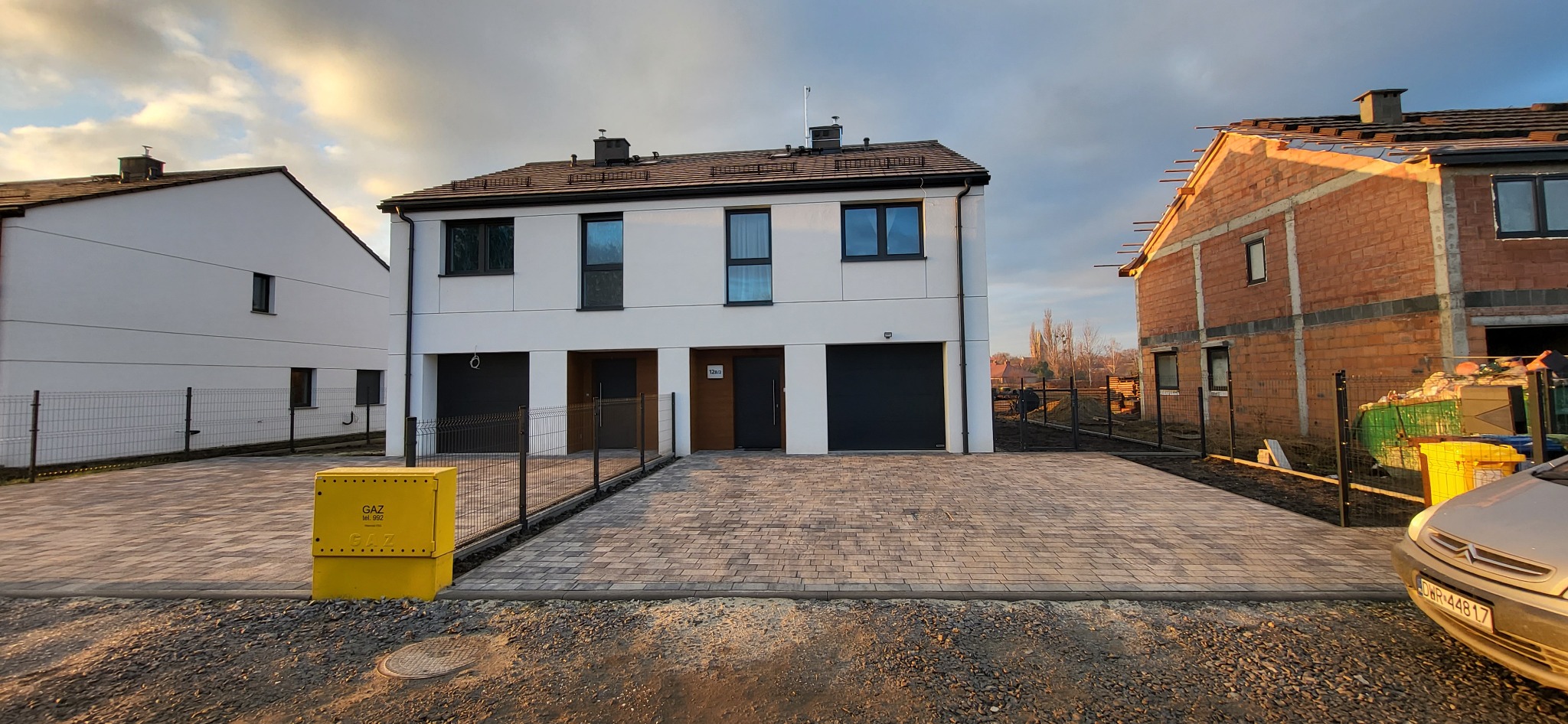 Nowoczesny dom dwukondygnacyjny z białymi panelami elewacyjnymi, szarą kostką brukową na podjeździe i żółtą skrzynką gazową, obok dom w trakcie budowy z czerwonej cegły.