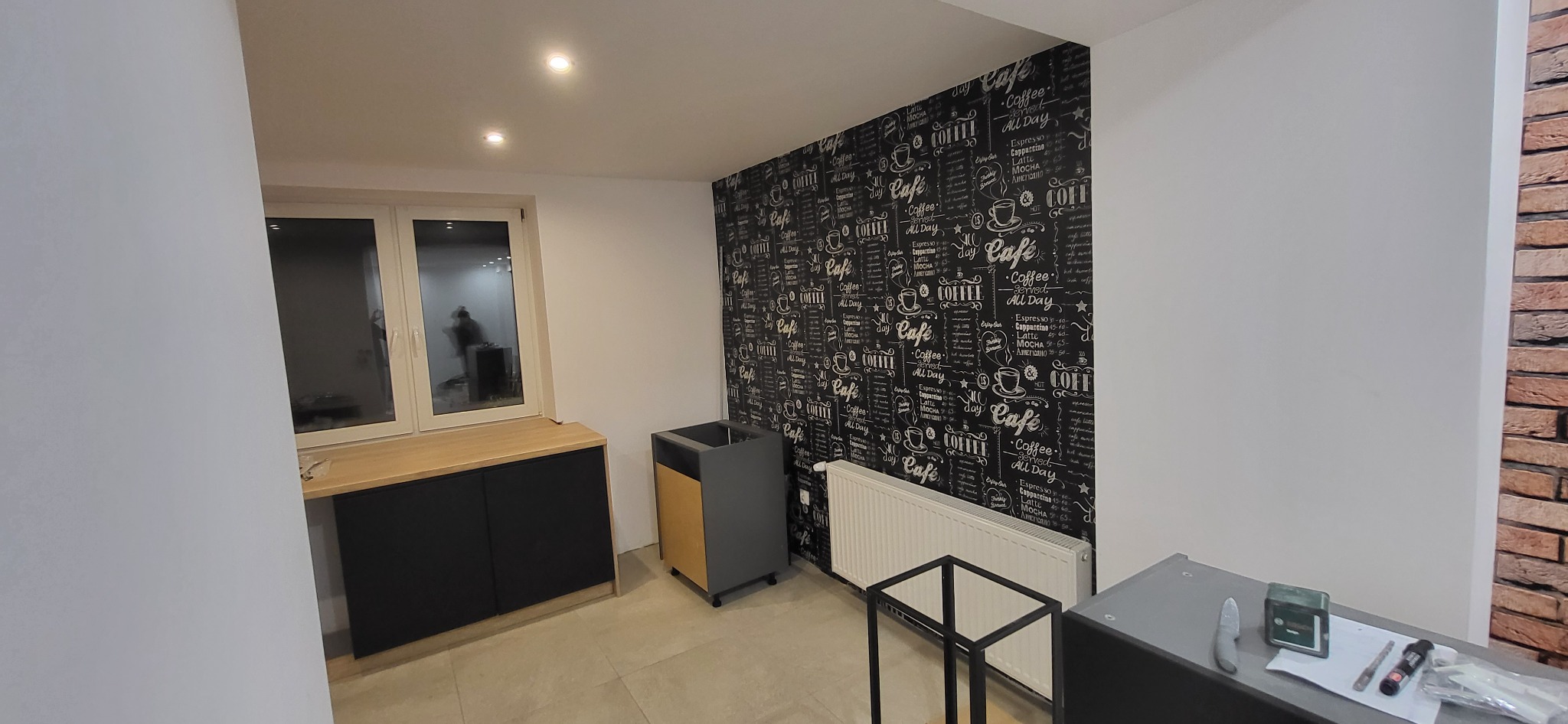 Wnętrze pokoju w trakcie wykańczania, z meblami kuchennymi w zabudowie, grzejnikiem i ścianą z tapetą w motywy kawowe, widoczne narzędzia na blacie.