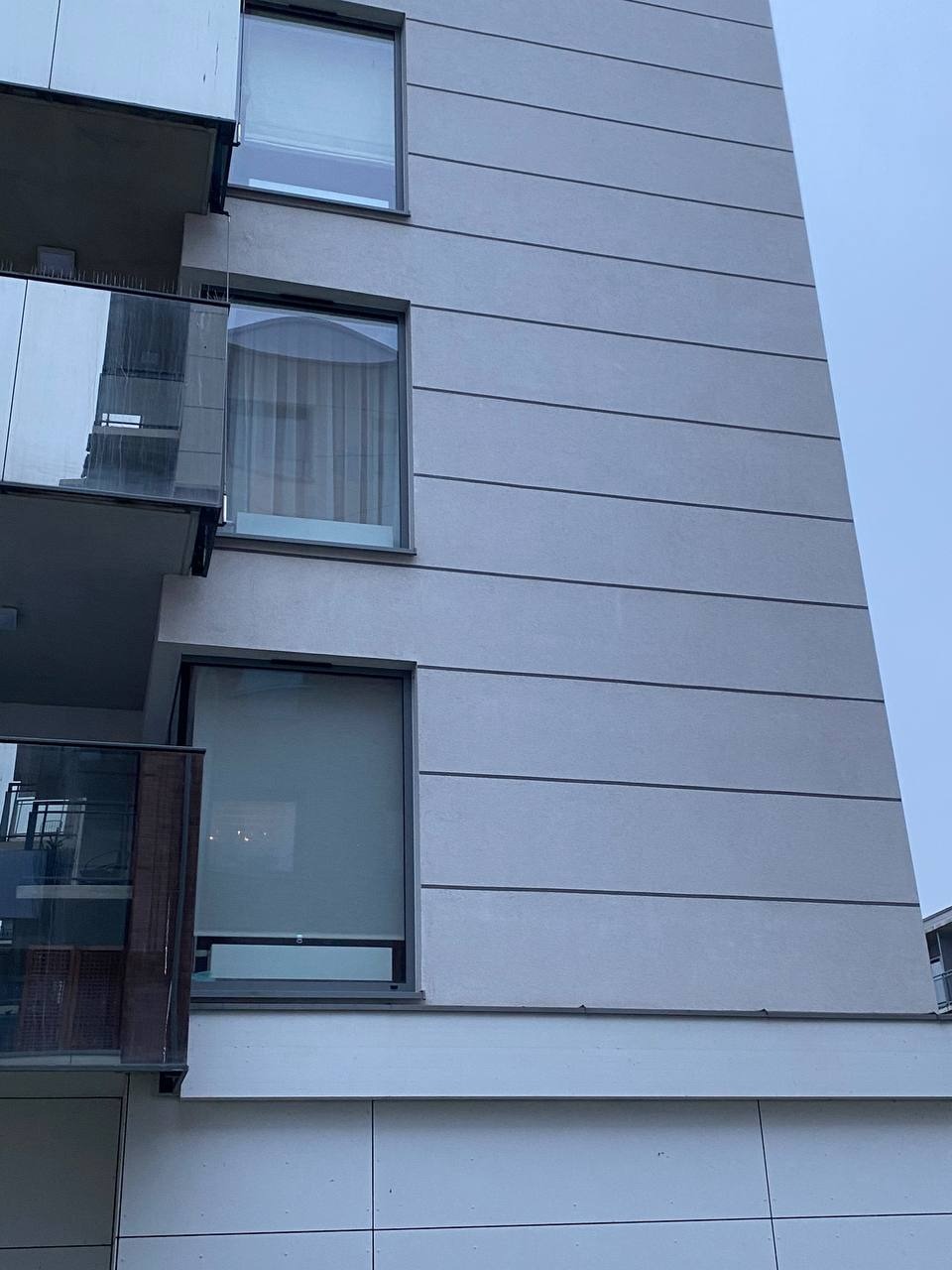 Fragment nowoczesnego budynku wielorodzinnego z szarą elewacją panelową i balkonami z przezroczystymi balustradami, widok z dołu.