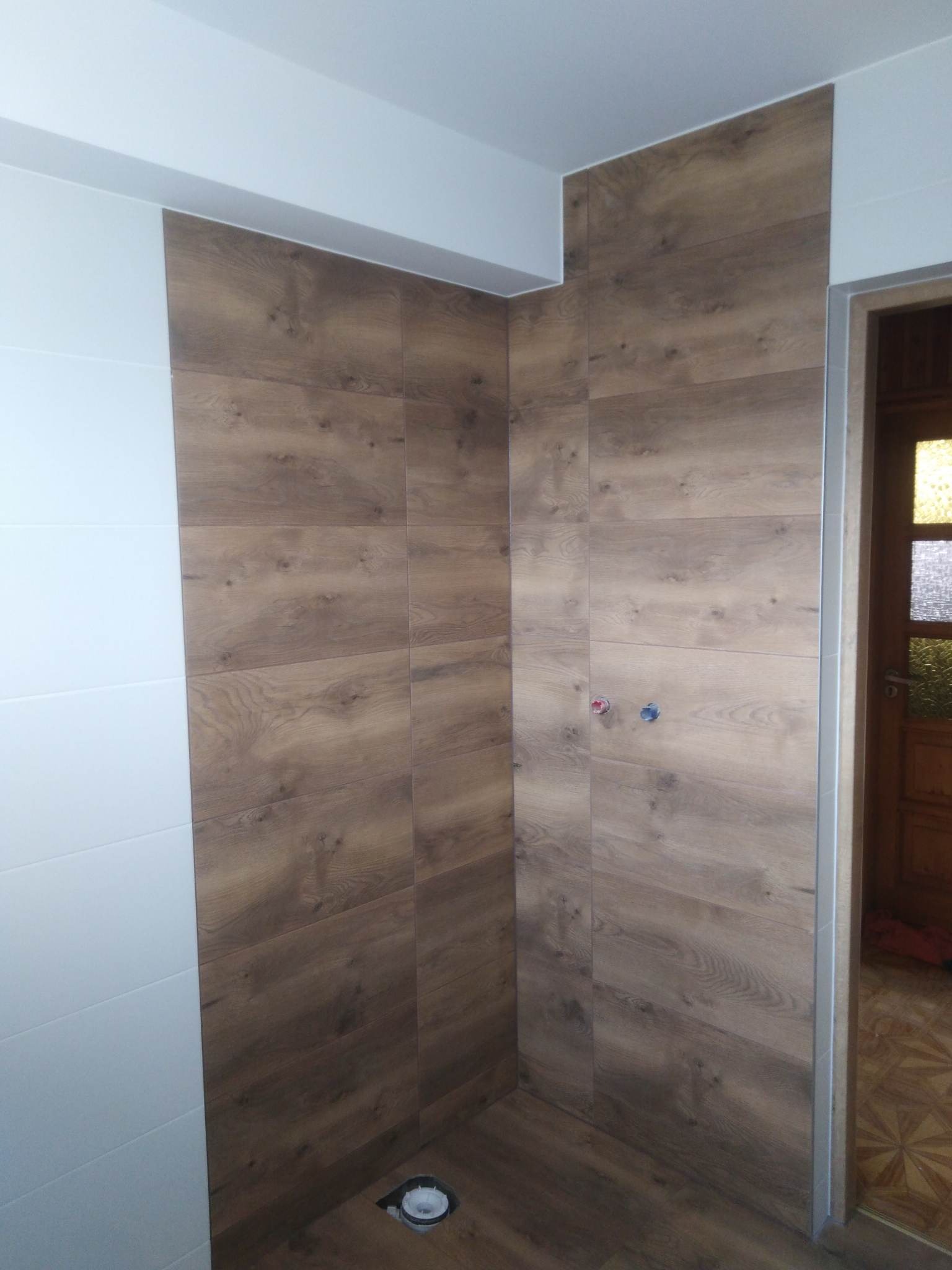 Ściany wyłożone płytkami imitującymi drewno w kabinie prysznicowej, widoczne przyłącza wody i odpływ podłogowy, obok drzwi z wzorzystą szybą.