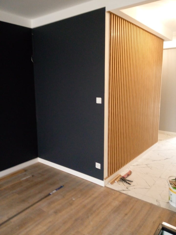 Ciemnogranatowa ściana połączona z drewnianą ścianką działową z listew, podłoga z paneli i marmurowych płytek, widoczne narzędzia i materiały wykończeniowe.