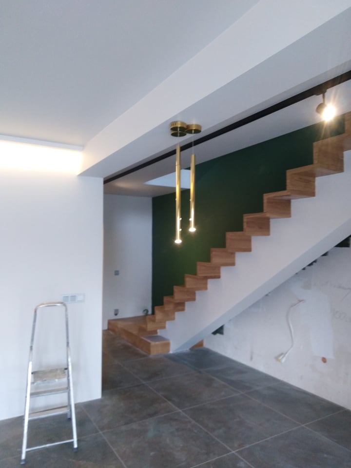 Wnętrze w trakcie prac wykończeniowych: drewniane schody z oświetleniem, ściana w kolorze butelkowej zieleni, szare płytki podłogowe, drabina malarska przy białej ścianie z widocznymi gniazdkami.