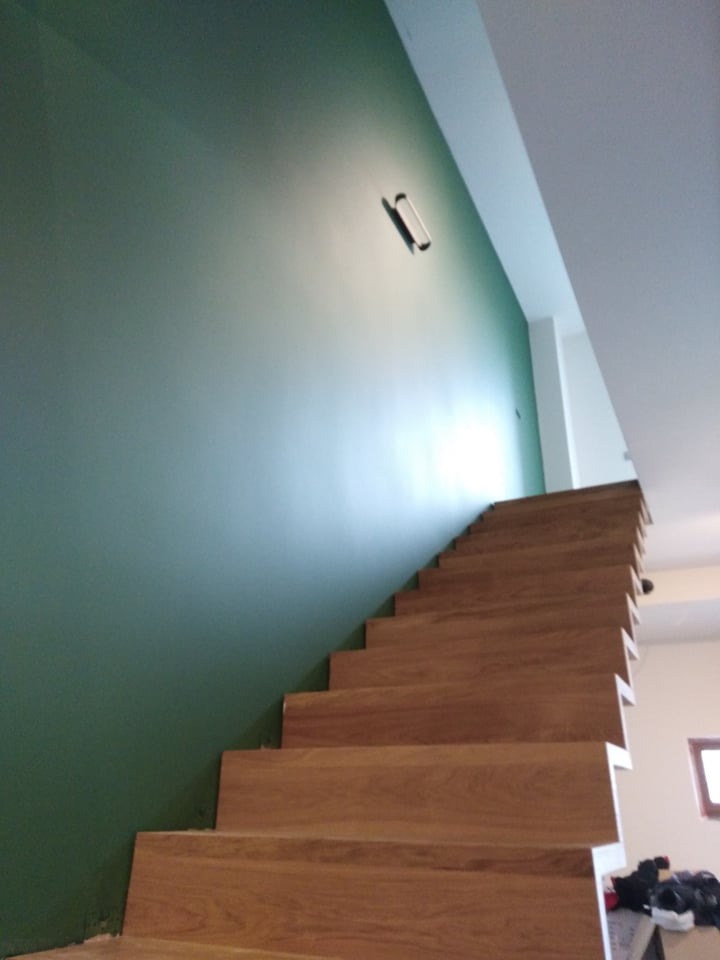 Ściana pomalowana na zielono z drewnianymi schodami, widoczna lampa ścienna i fragment sufitu.