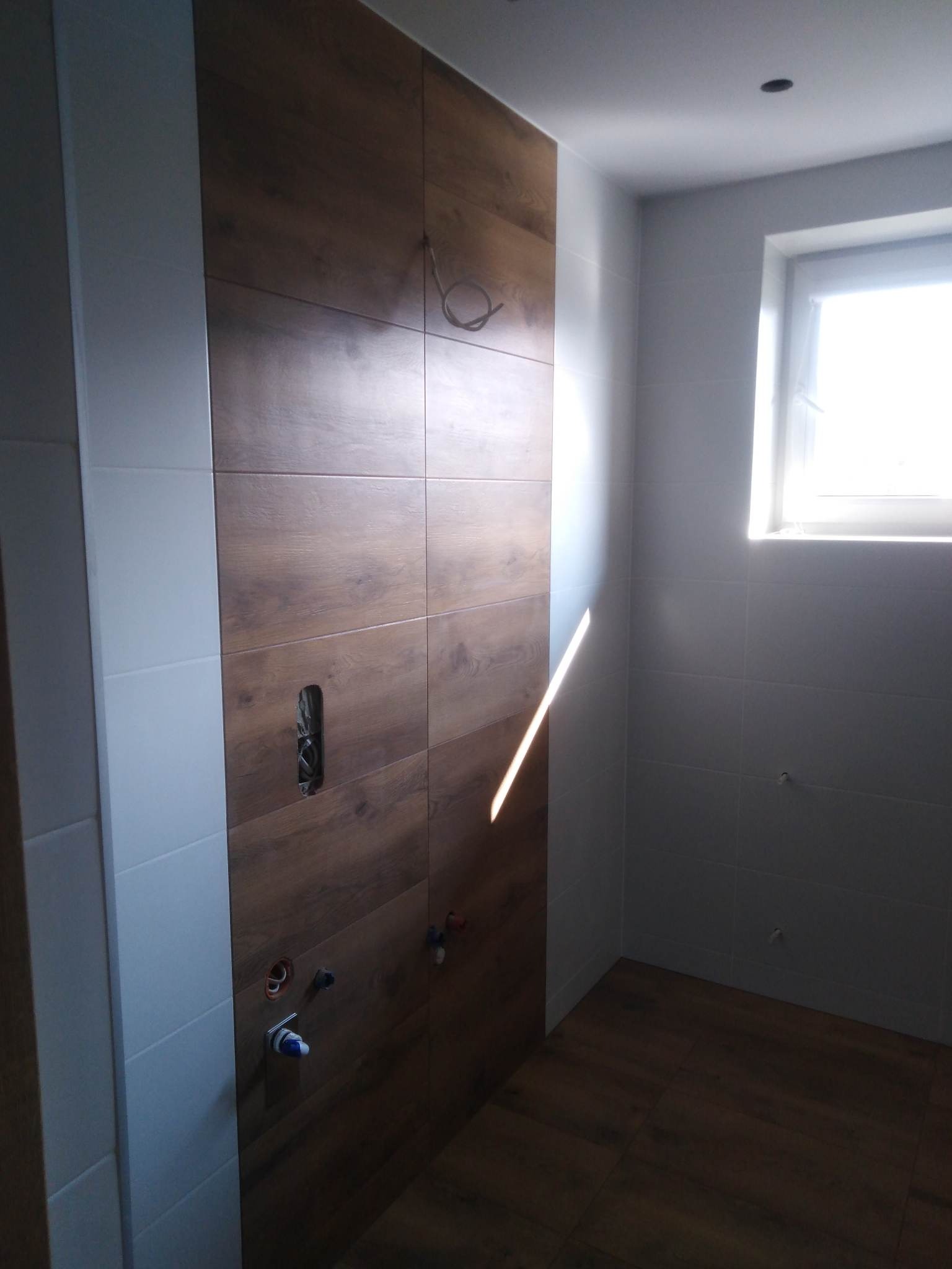 Wykończona łazienka z drewnopodobnymi płytkami na ścianie i podłodze, widoczne przygotowane instalacje wodne i elektryczne, oraz fragment okna z silnym światłem słonecznym.