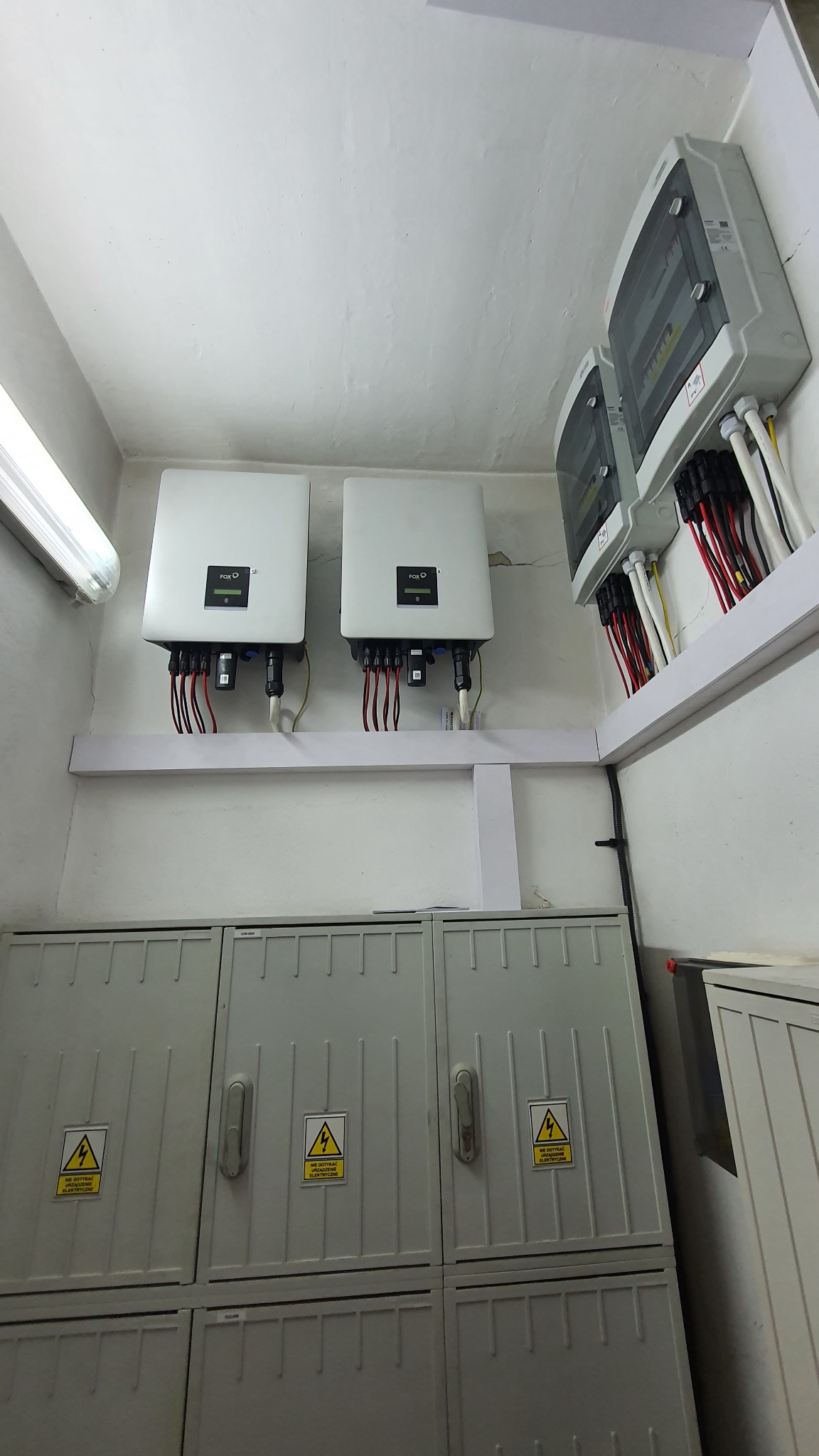 Dwa inwertery marki FOX-O zamontowane na ścianie nad szarą szafą elektryczną z ostrzeżeniami o wysokim napięciu. Dodatkowe skrzynki elektryczne widoczne po prawej stronie.