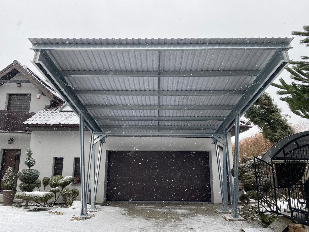 Zadaszenie na samochód ze srebrnej blachy falistej na metalowej konstrukcji podczas opadów śniegu, na tle domu z białym tynkiem i brązowym dachem.