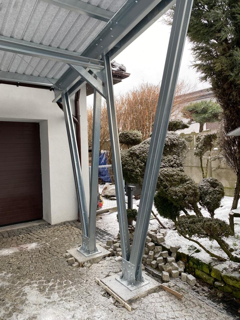 Stalowa konstrukcja z zadaszeniem, prawdopodobnie przygotowana pod montaż paneli słonecznych, widoczna na tle budynku z brązową bramą garażową i zaśnieżonego ogrodu z ozdobnymi krzewami.