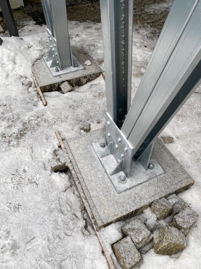 Metalowa konstrukcja wsporcza na betonowej podstawie, częściowo przysypana śniegiem, z widocznymi elementami montażowymi i kostką brukową w tle.