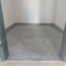 Szare płytki podłogowe ułożone w małym pomieszczeniu z białymi ścianami i szarymi listwami przypodłogowymi, widoczne fragmenty ościeżnicy drzwiowej.