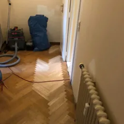 Fragment podłogi w trakcie układania parkietu w jodełkę, widoczny odkurzacz przemysłowy Starmix, zabezpieczona ściana i grzejnik.