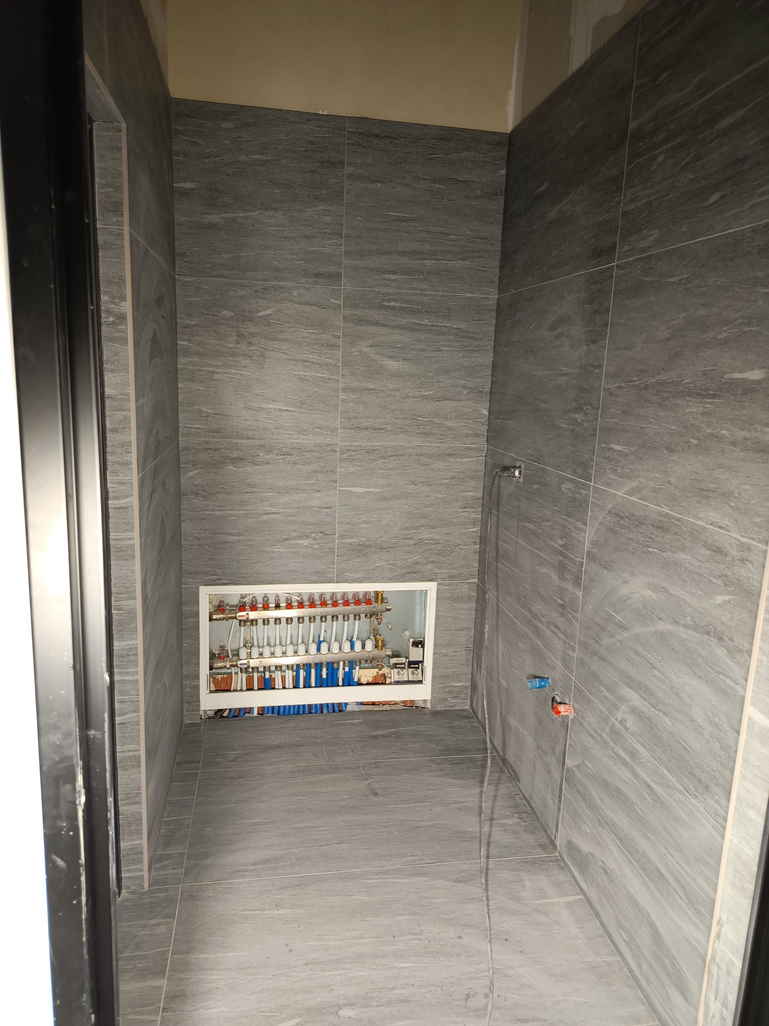 Szara łazienka w trakcie remontu z widoczną rozdzielnicą ogrzewania podłogowego i instalacją wodną, płytki imitujące kamień na ścianach i podłodze, surowy stan wykończenia.