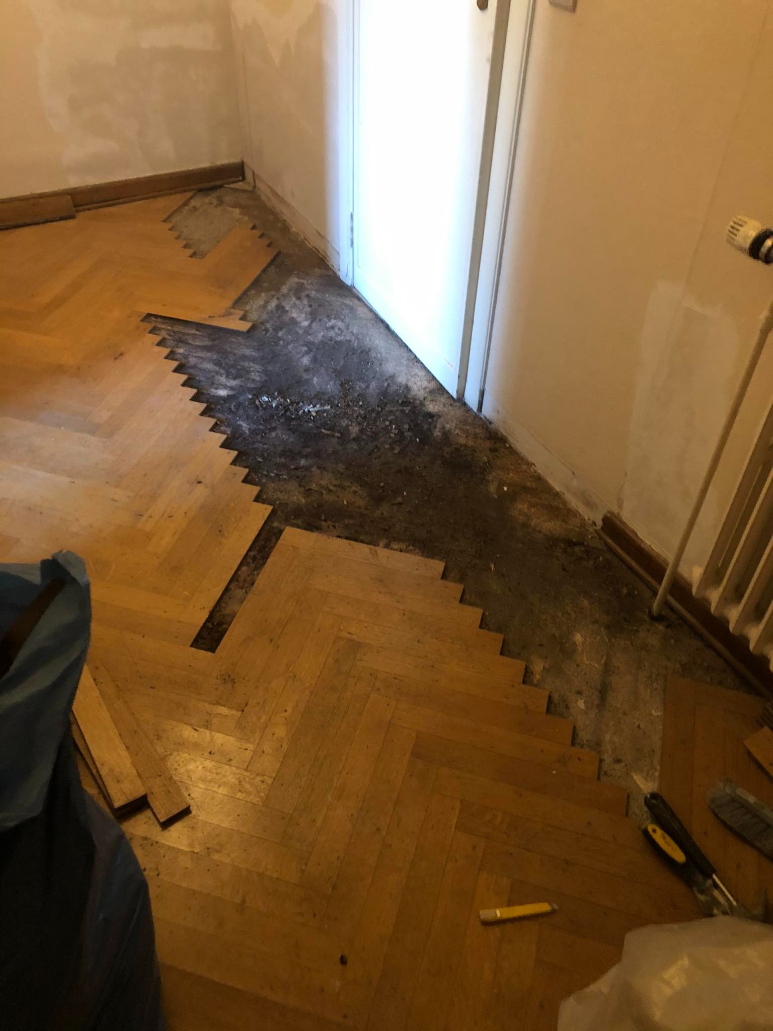 Fragment pokoju z częściowo usuniętą starą parkietową podłogą w jodełkę, odsłaniającą warstwę podkładową. Widoczne narzędzia, worek na odpady i grzejnik.
