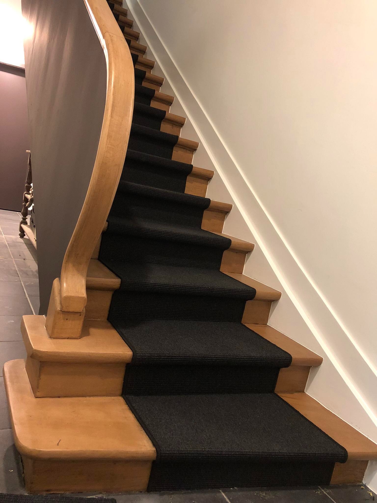 Schody drewniane z czarną wykładziną dywanową, jasne drewno balustrady, ściana pomalowana na biało, fragment podłogi z szarych płytek.