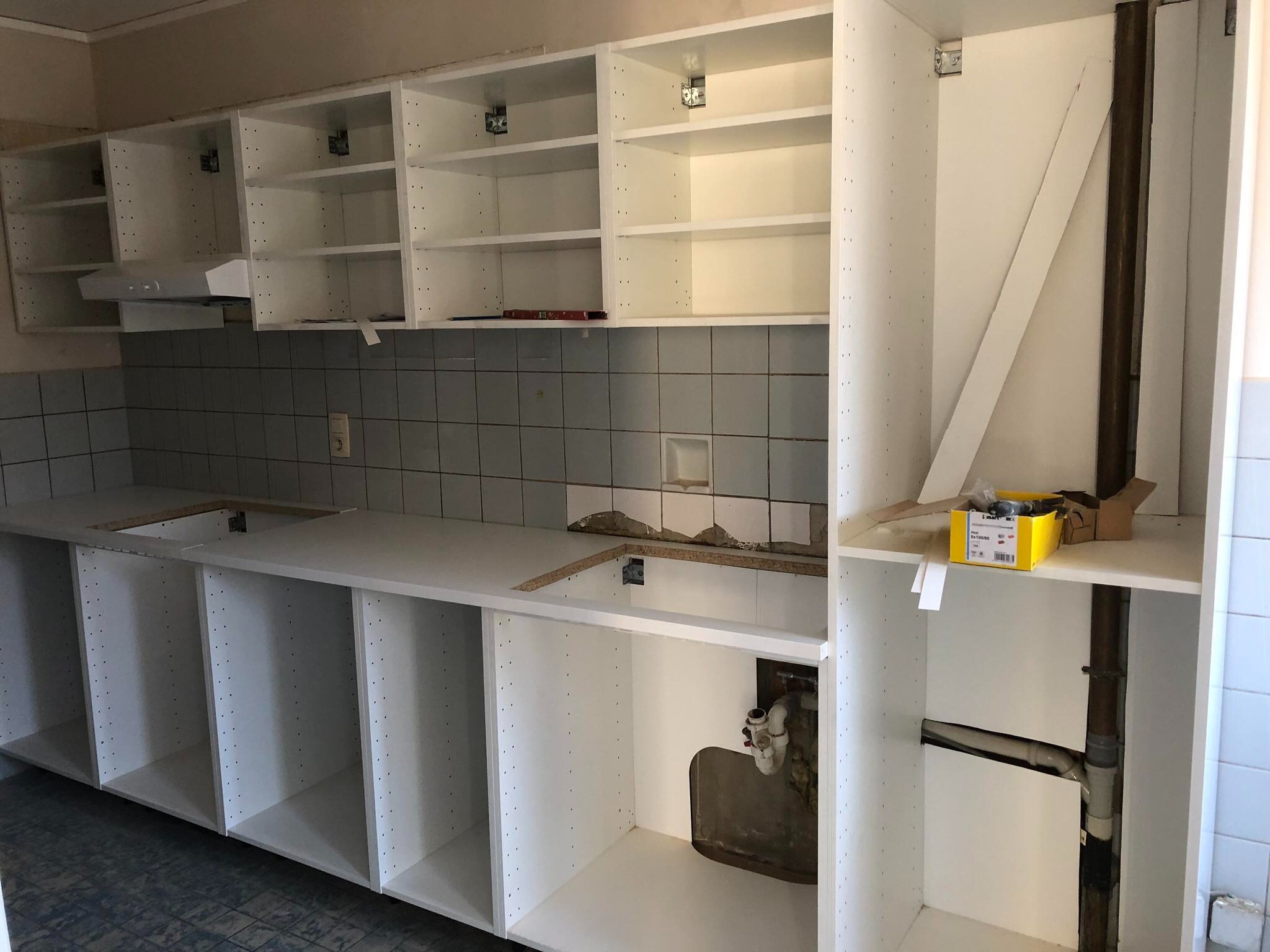 Wnętrze kuchni w trakcie montażu białych szafek kuchennych, widoczne szare płytki na ścianie i niezabezpieczone rury instalacyjne.