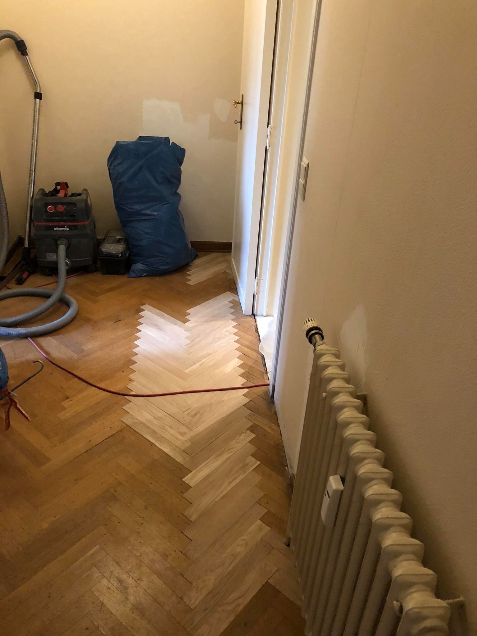 Fragment podłogi w trakcie układania parkietu w jodełkę, widoczny odkurzacz przemysłowy Starmix, zabezpieczona ściana i grzejnik.