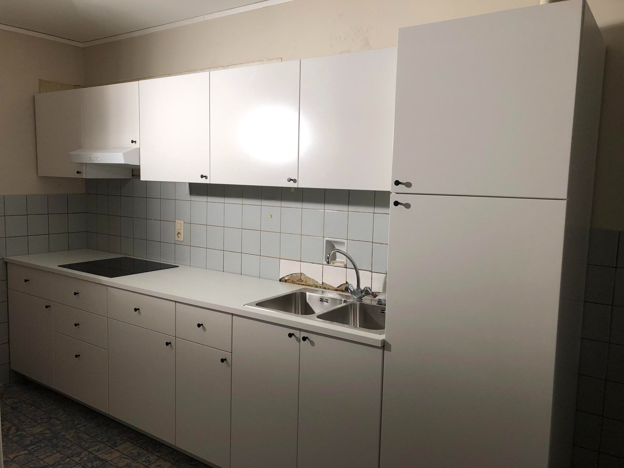 Kuchnia przed remontem: Białe szafki kuchenne z czarnymi uchwytami, dwukomorowy zlew ze srebrną baterią, płyta indukcyjna, okap, jasne kafelki na ścianie, uszkodzone kafelki nad zlewem.