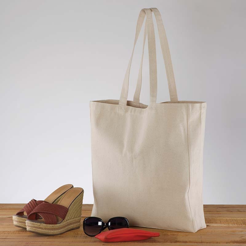 Beżowa torba płócienna typu tote bag z długimi uchwytami, stojąca na drewnianym blacie obok brązowych sandałów na koturnie, okularów przeciwsłonecznych i czerwonego etui.