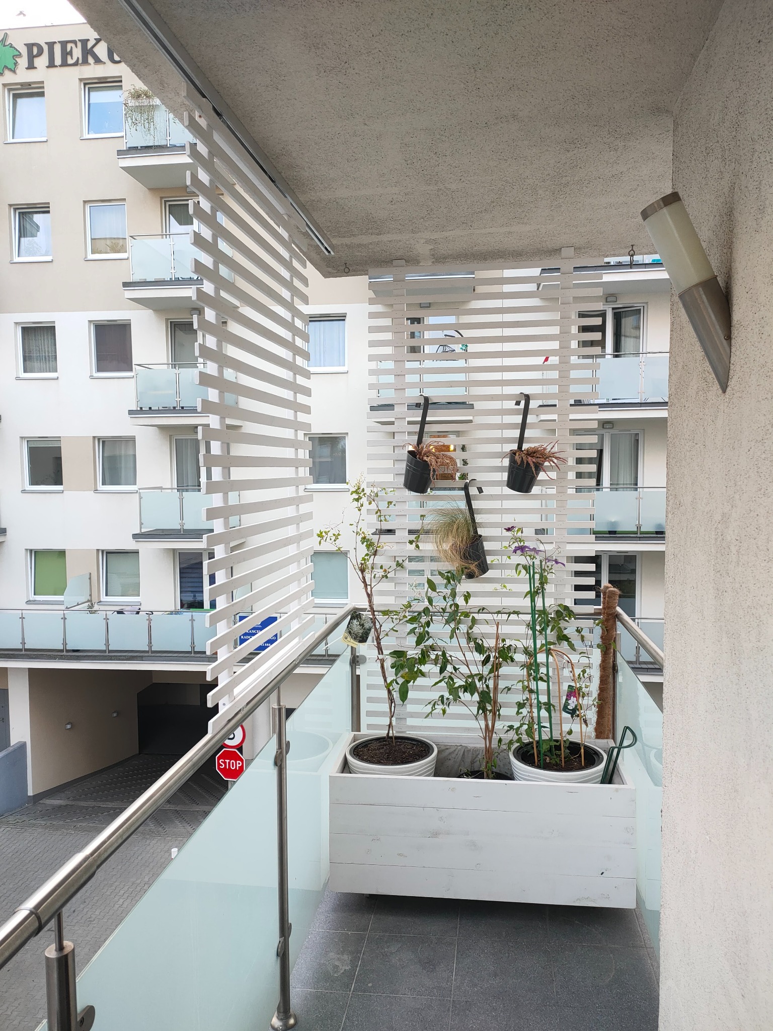 Zabudowany balkon z białymi lamelami, drewnianą skrzynią z roślinami i wiszącymi doniczkami, widok na sąsiedni budynek.