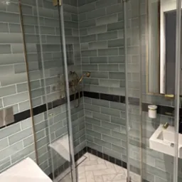 Nowoczesna kabina prysznicowa ze złotymi detalami w Niebocku, z widoczną toaletą i umywalką w aranżacji łazienki z szaro-błękitnymi płytkami.