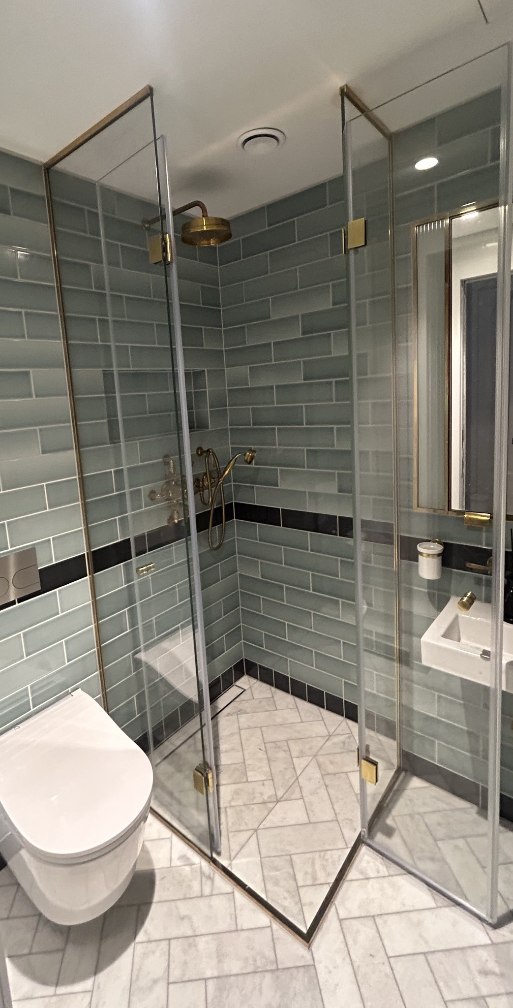 Nowoczesna kabina prysznicowa ze złotymi detalami w Niebocku, z widoczną toaletą i umywalką w aranżacji łazienki z szaro-błękitnymi płytkami.