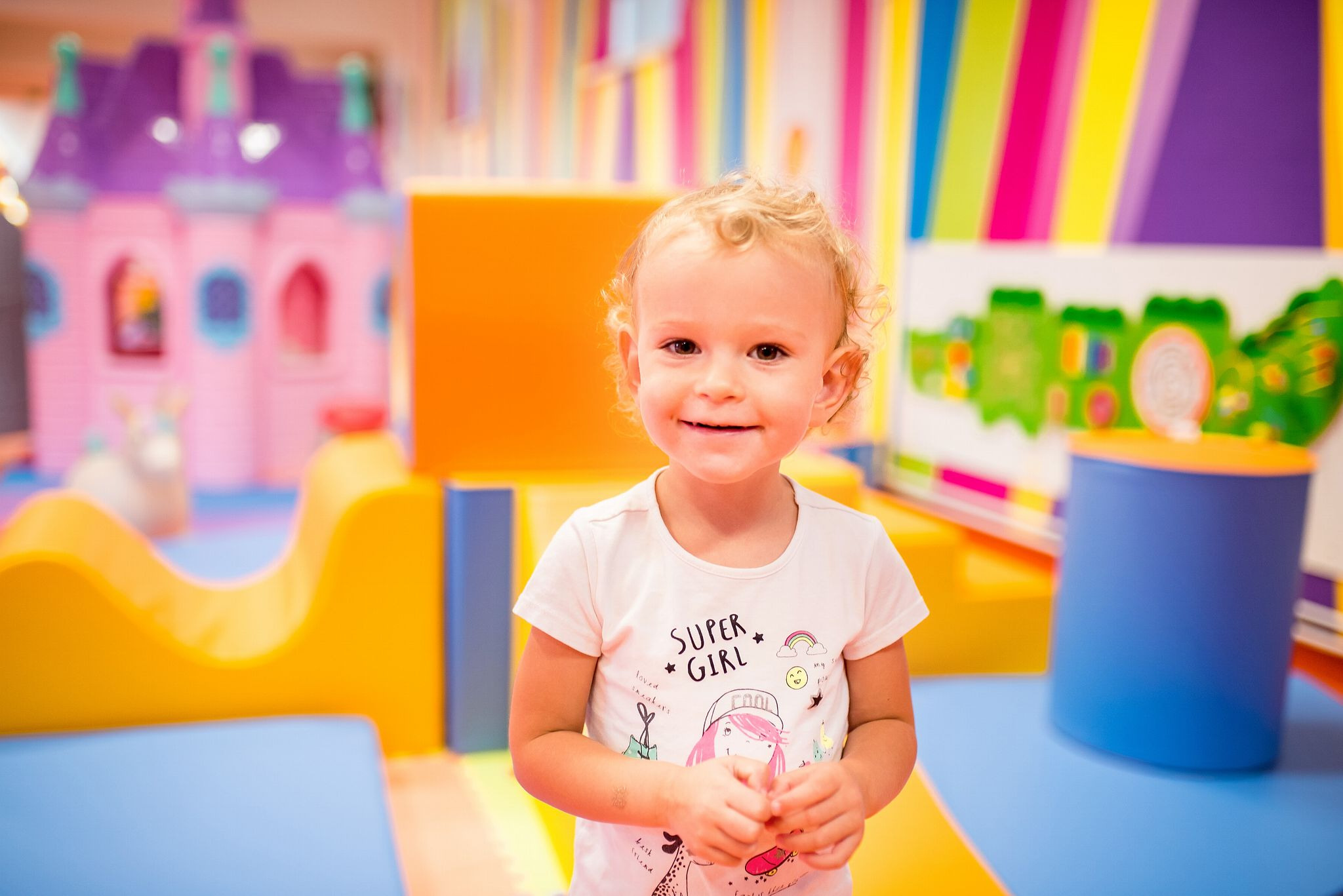 Uśmiechnięta dziewczynka w koszulce z napisem 'Super Girl' na tle kolorowej strefy zabaw dla dzieci z miękkimi elementami i zamkiem księżniczki.