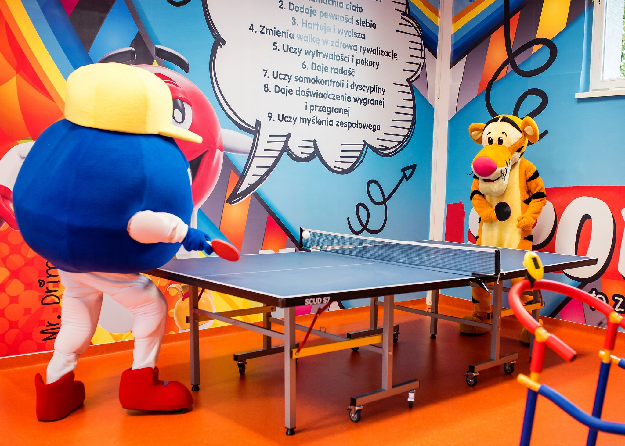 Dwaj animatorzy w kostiumach, jeden w stroju niebieskiego stworka z żółtą czapką gra w ping ponga, drugi w przebraniu Tygryska obserwuje grę, w tle kolorowa ściana z napisem.