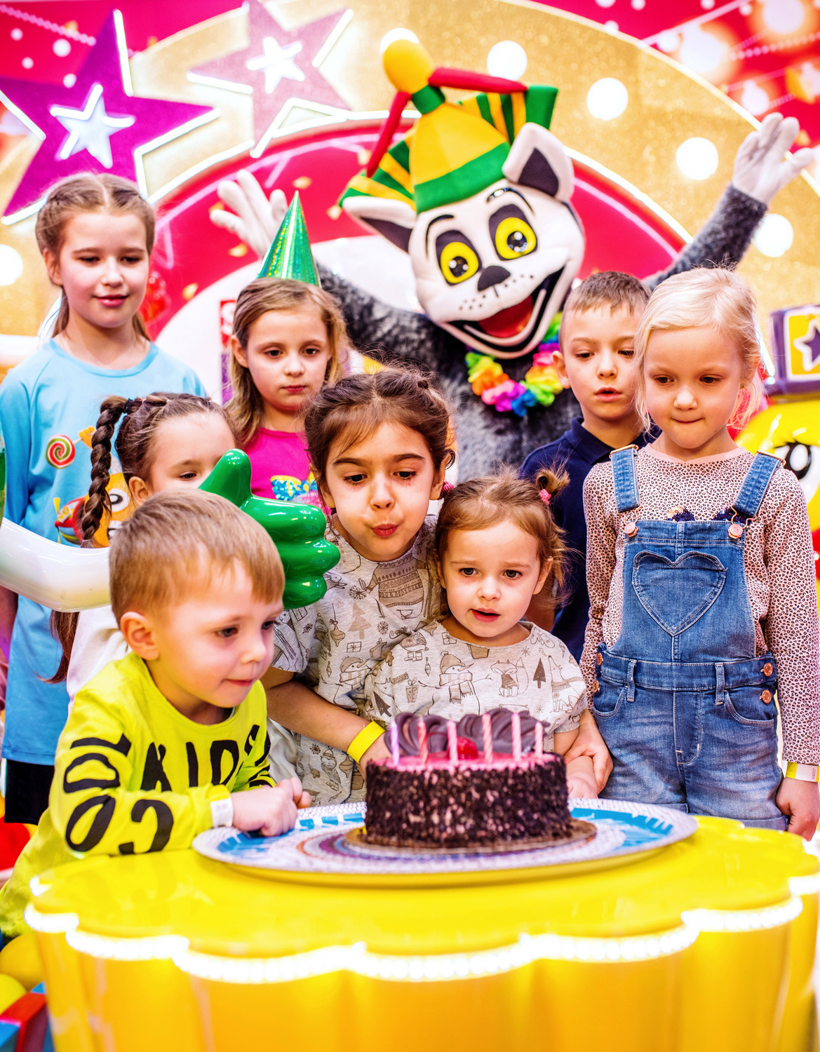 Grupa dzieci zgromadzona wokół tortu urodzinowego, za nimi postać w kostiumie kota z kolorową czapką i girlandą na szyi, w tle dekoracje z gwiazdami i jasne oświetlenie.