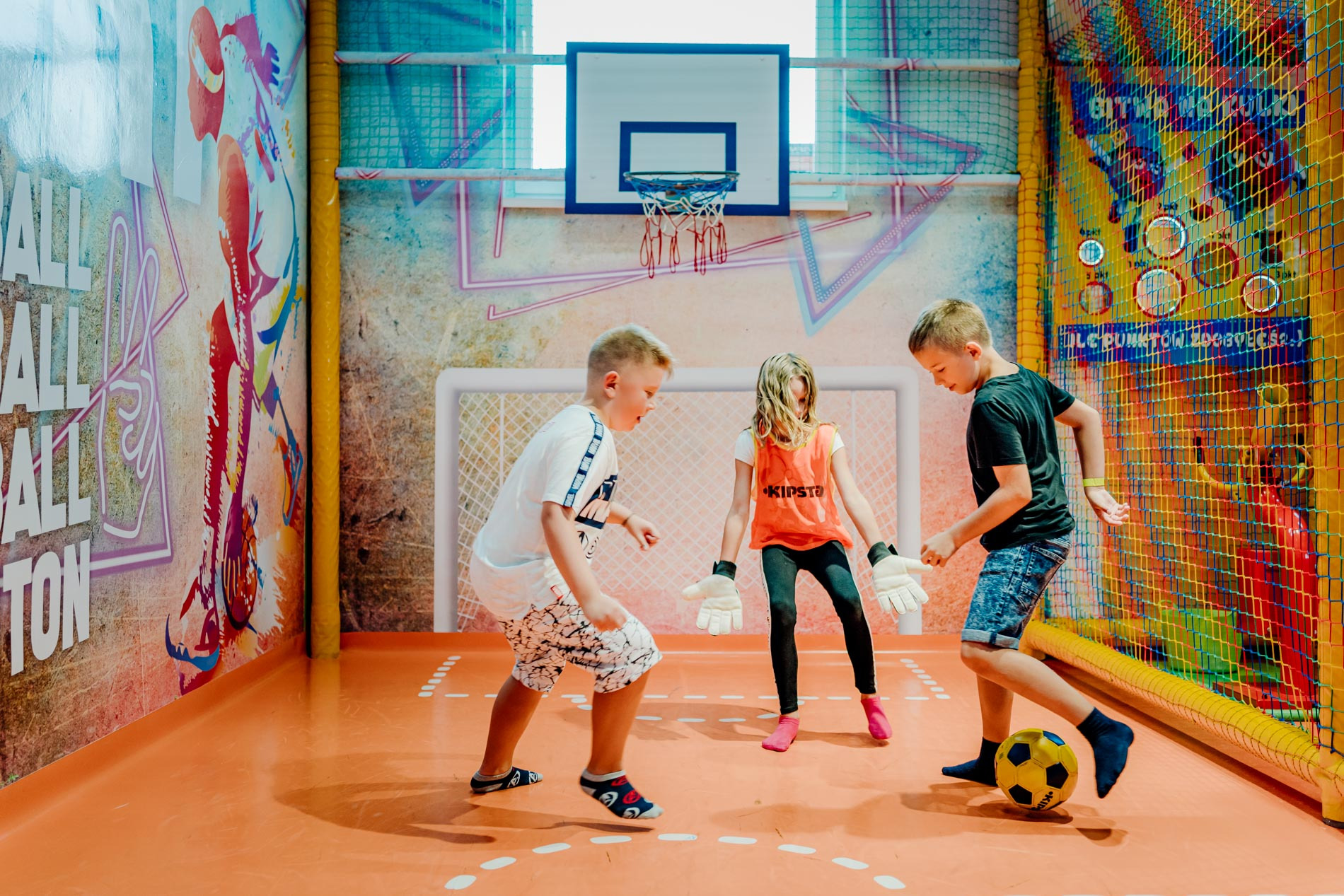 Trójka dzieci w różnym wieku gra w piłkę nożną na pomarańczowym boisku w hali sportowej z koszem do koszykówki, bramką do piłki nożnej i kolorową siatką zabezpieczającą.