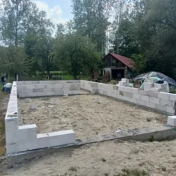Wznoszenie fundamentów z bloczków betonowych na piaszczystym gruncie, widoczny pracownik z taczką w tle, drewniany budynek gospodarczy i drzewa.