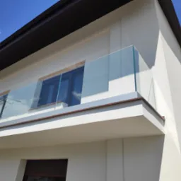 Nowoczesny balkon z przezroczystą taflą szkła, zabezpieczającą przestrzeń, z widocznym odbiciem nieba i instalacji elektrycznej w szybie, na tle jasnej elewacji budynku i błękitnego nieba.