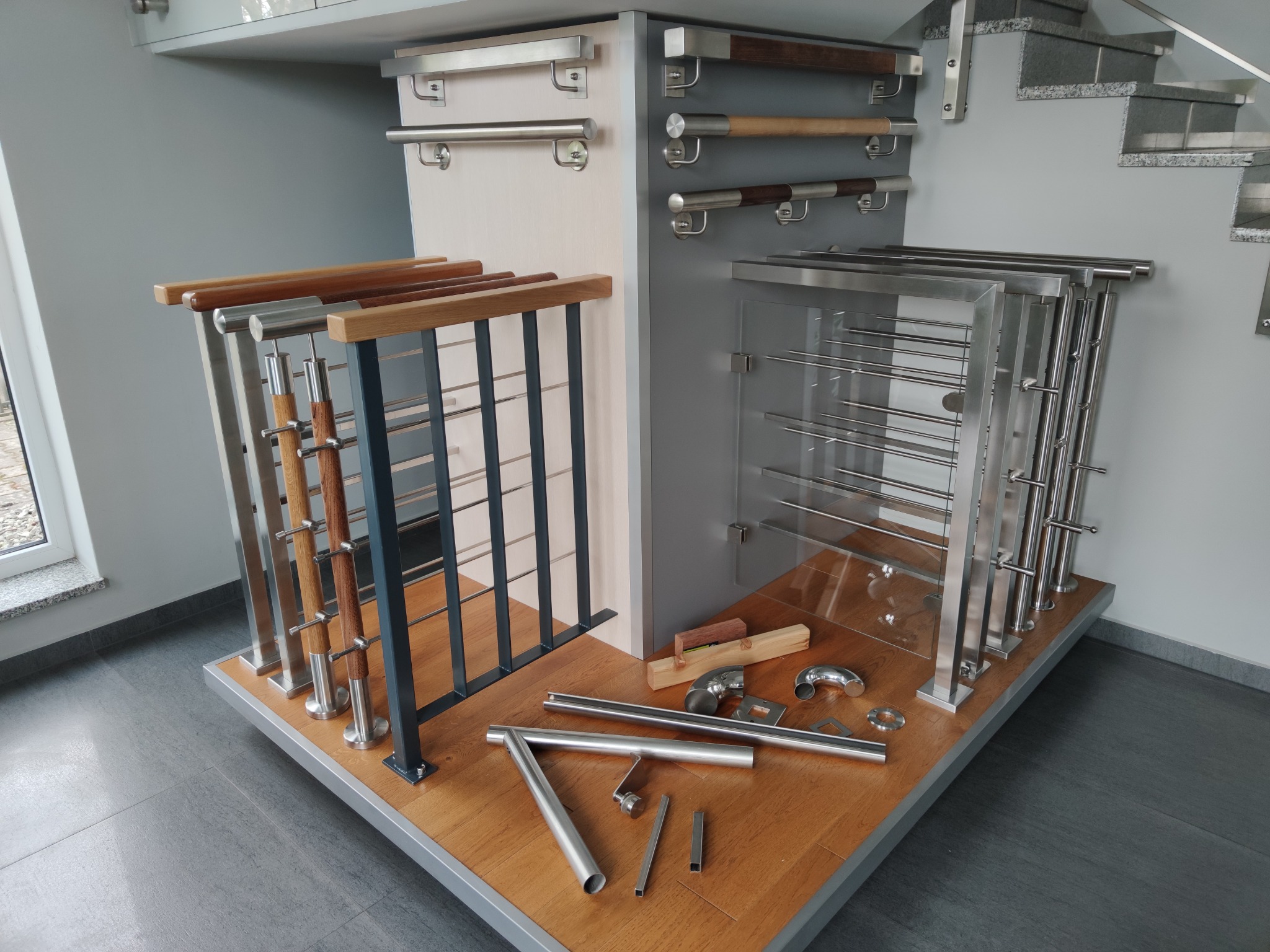 Ekspozycja różnych modeli balustrad i poręczy: nierdzewne, szklane i z elementami drewnianymi, prezentowane w showroomie, obok schodów z szarymi stopniami.