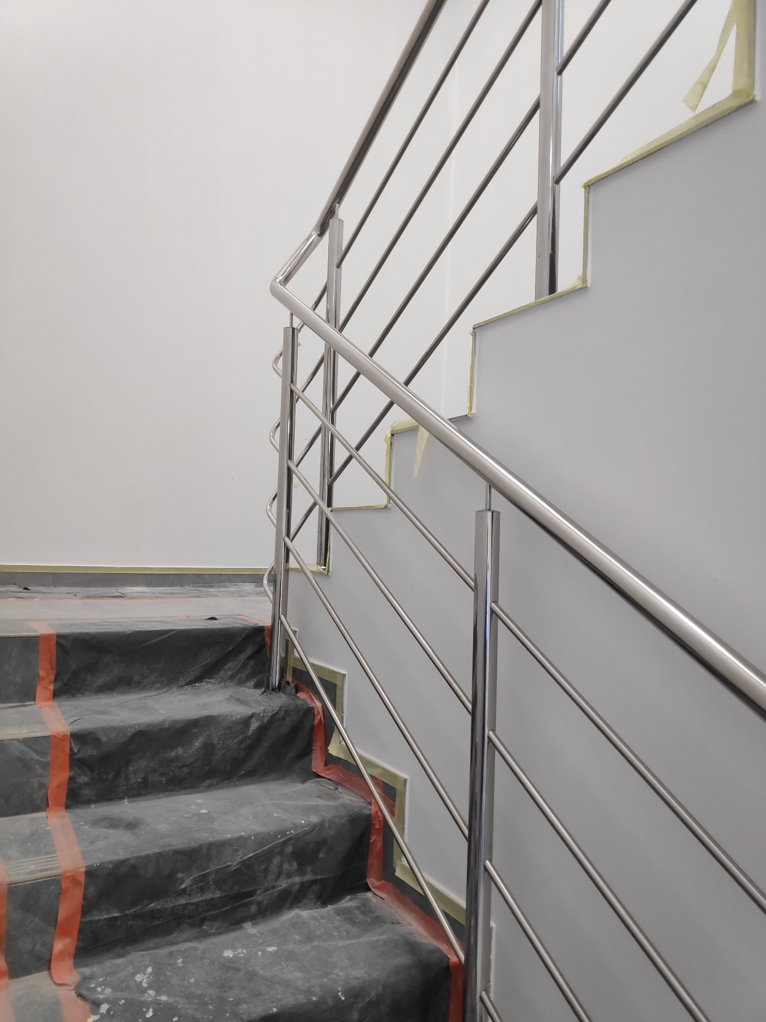 Montaż stalowej poręczy na schodach, zabezpieczonych folią malarską i taśmą, widoczne ślady prac wykończeniowych.
